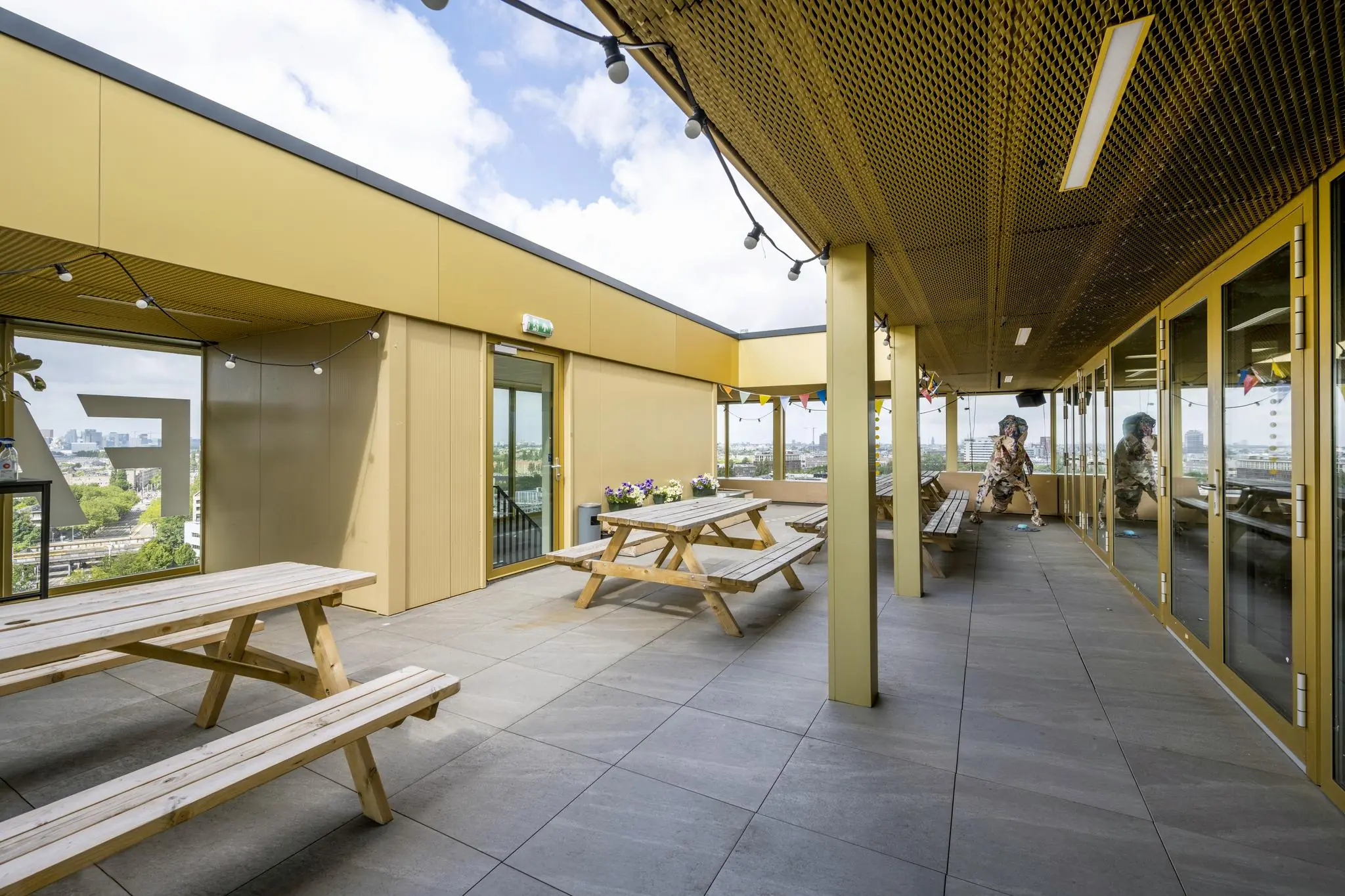 Dakterras aan de James Wattstraat met houten picknicktafels, gele gevels en uitzicht op de stad.